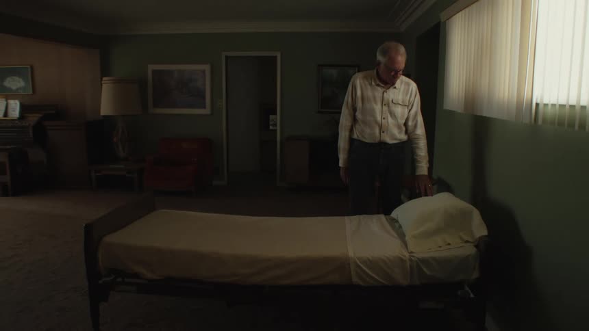 Elderly person beside a bed in a room with a sentimental expression