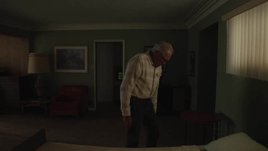 A man walking around the dark room making the bed