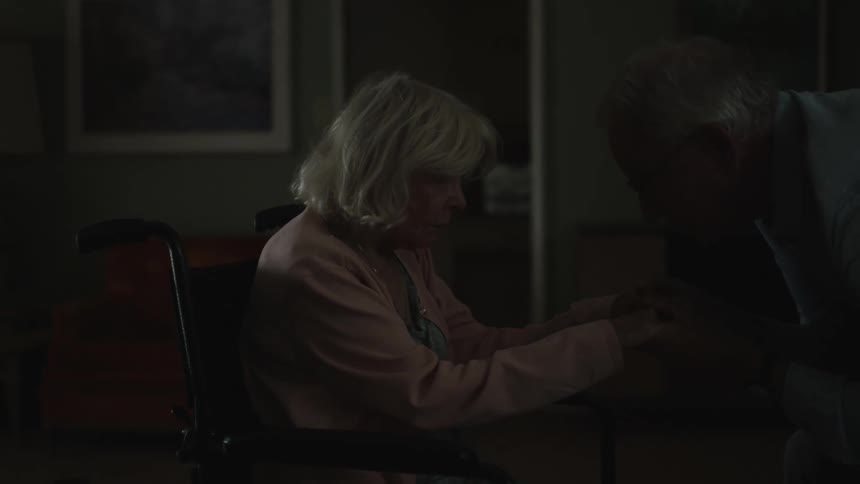 Elderly person leading his wife into the room and assisting her in sitting on the bed
