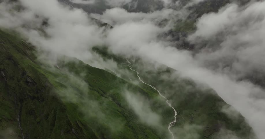 An aerial view of a valley between mountains covered in vegetation and lots of misty clouds overhead