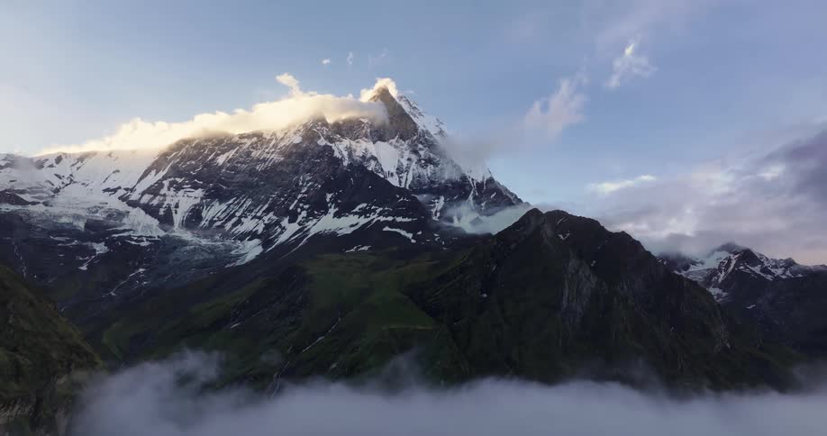 Snow covered mountain towers over smaller green mountains covered by low lying clouds