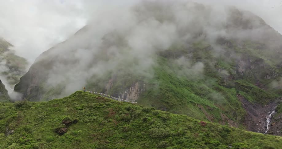A person standing on top of a grassy hill and behind a huge mountain with a lot of fog