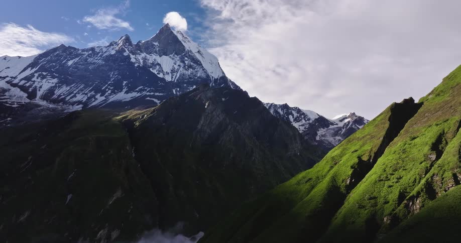 Tall snow covered mountain towers over smaller green mountains on a cloudy day