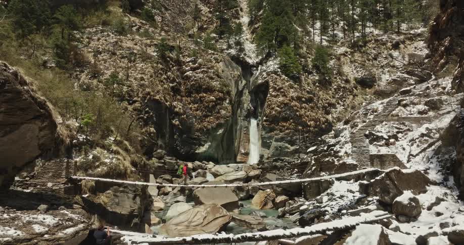 Woman crossing a narrow bridge in a natural environment with rocks and a waterfall