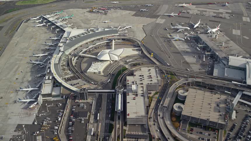 Aerial of roads winding around the terminal of an airport with planes lined up around the loading gates near a runway
