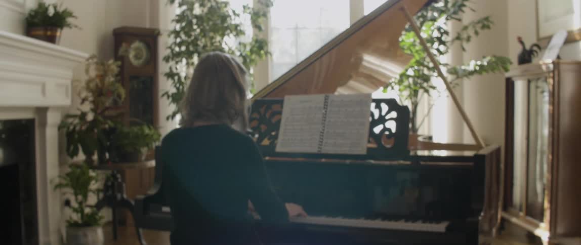 A woman has plants around her piano as she plays