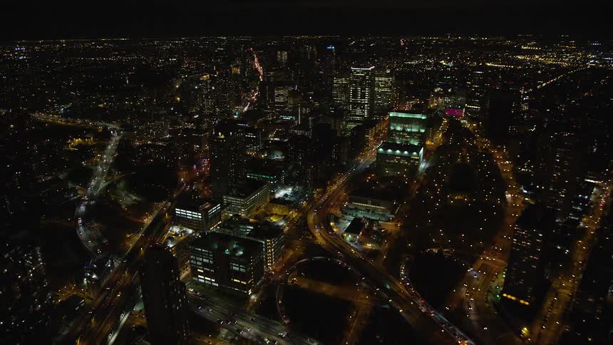 Aerial over a lit up bridge next to a metropolitan city where the headlights of vehicles move along the roads between tall buildings at night