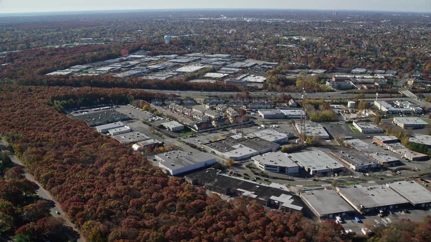 Traffic flows along highway past business complexes and residential neighborhoods separated by woods with fall foliage
