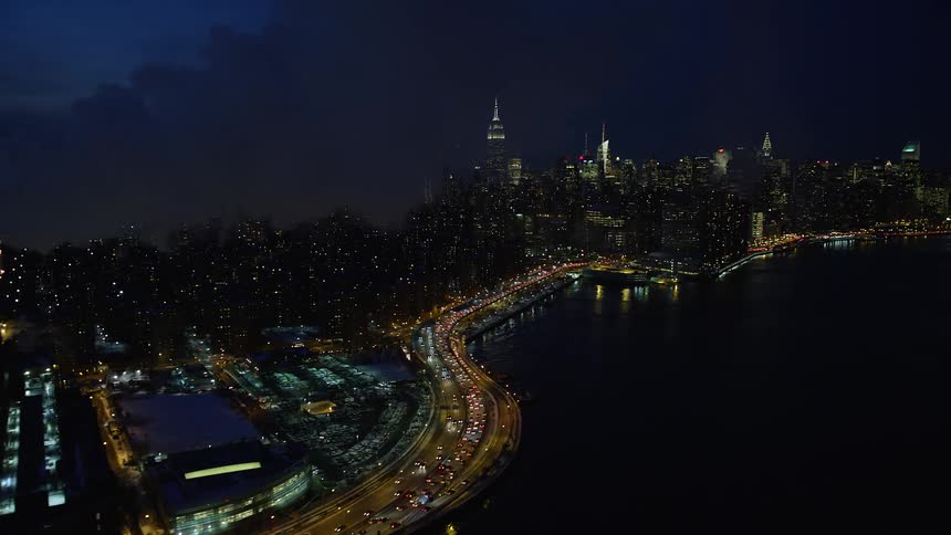 Aerial of a metropolis shoreline at night passing by a cloud of smoke coming from a factory blowing out over the water