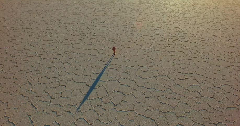 The sun shines upon a person walking across the baren dry cracked desert ground casting a shadow behind them as they walk