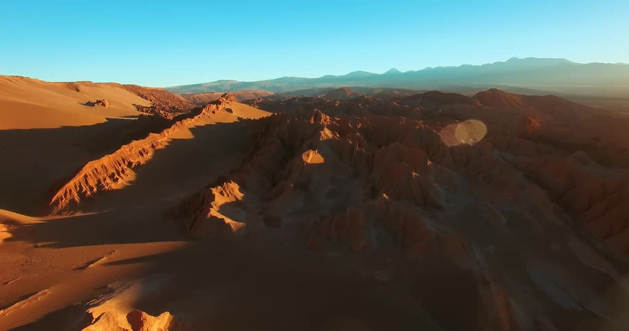 The sun shines upon desert mountains and rock formations under a clear blue sky