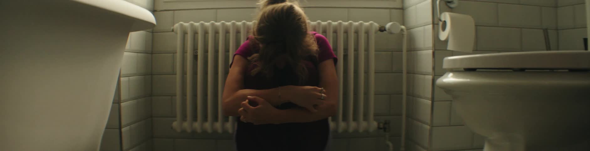Woman sits huddled on bathroom floor near radiator