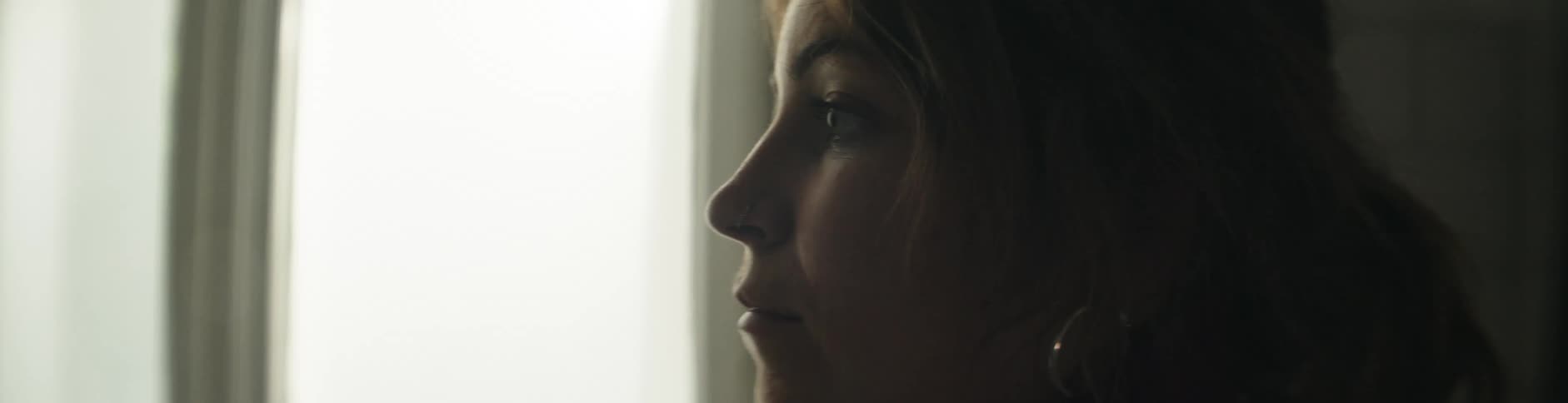 A woman wearing earrings and a ponytail gazes into space and then turns her attention towards the window