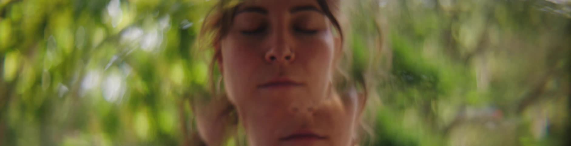 Woman with brown hair does yoga in front of green trees