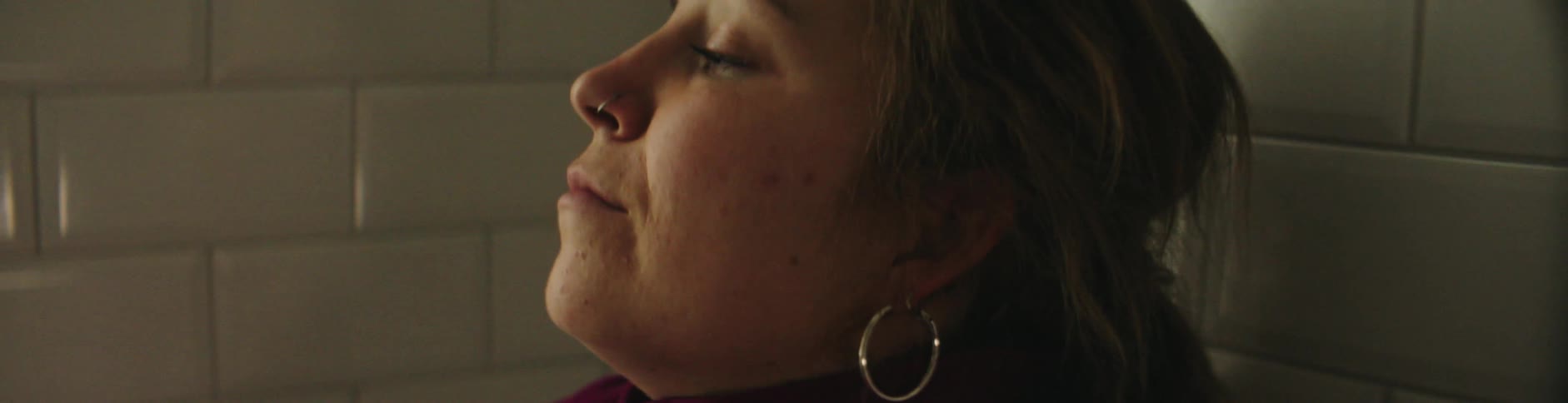 Woman with hoop earrings piercing and a red shirt leans her head against the wall in the bathroom