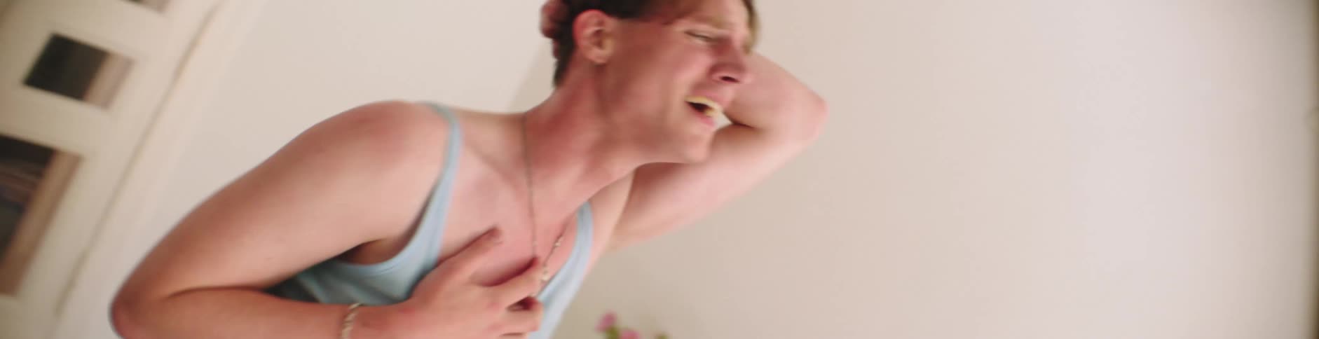 A man donned in a light blue tank top and a necklace dances with one hand gracefully placed on the back of his head