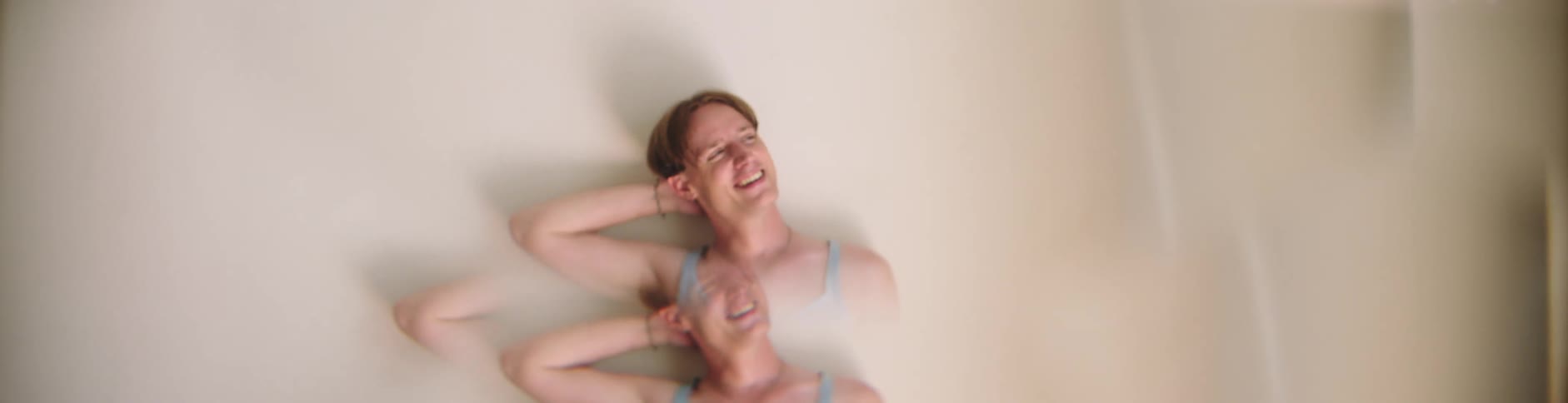 Man with a necklace and a light blue tank top leans against the wall laughing