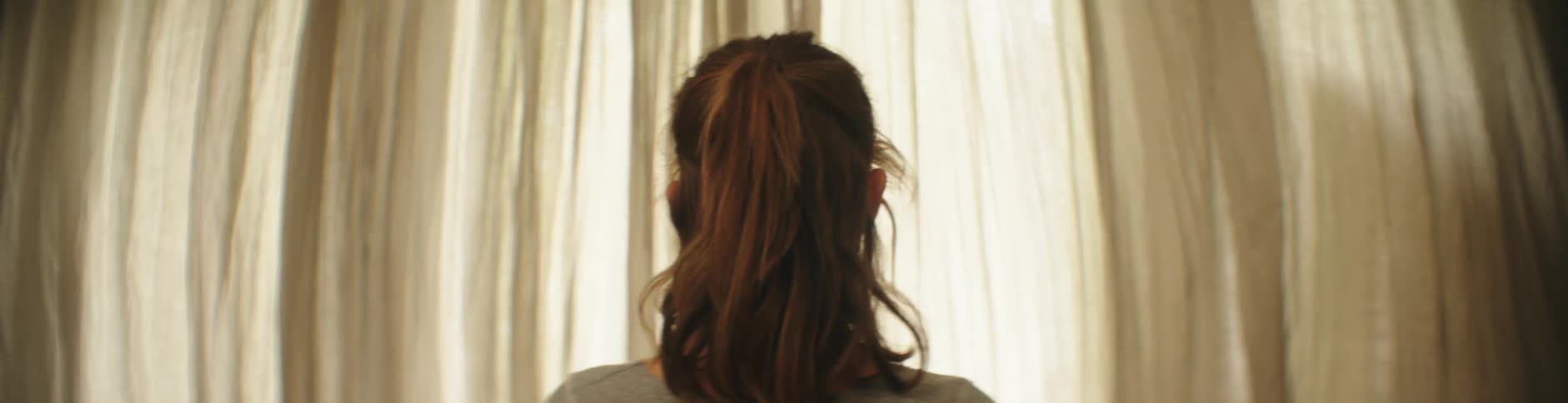 A lady alks to the window and spread the curtain apart to have a view of outdoor through the window