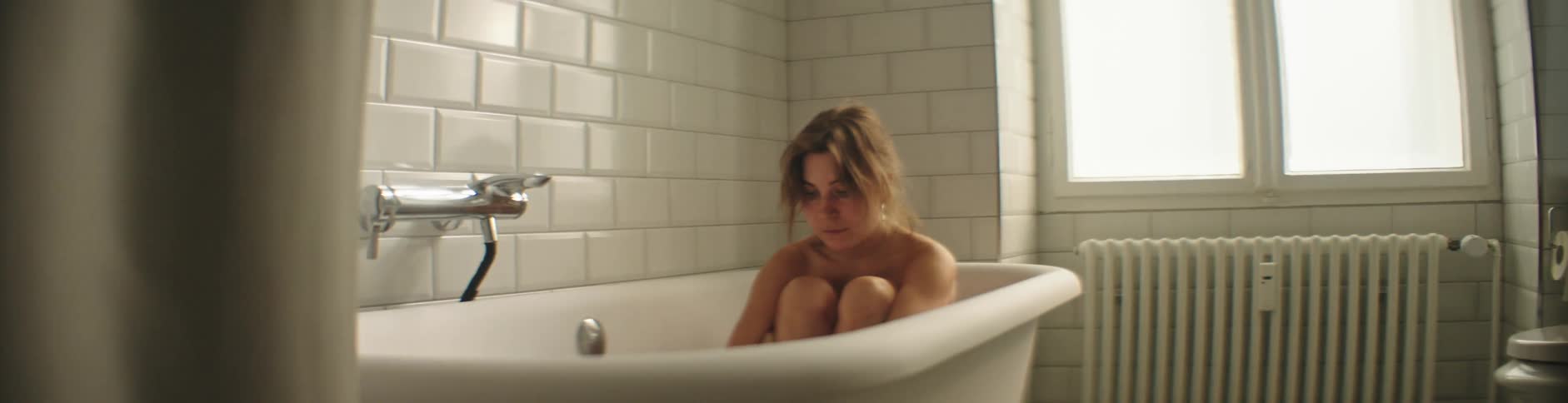 Woman sits huddled in tub in white bathroom