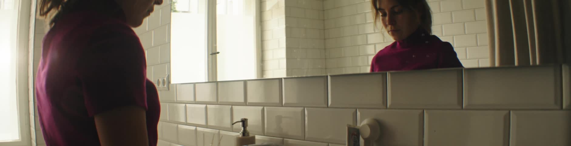 A woman with brown hair and a red shirt stands in front of the bathroom mirror looking down contemplatively