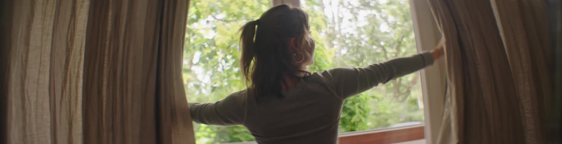 A woman opening a curtain from a window that sees some tree leaves