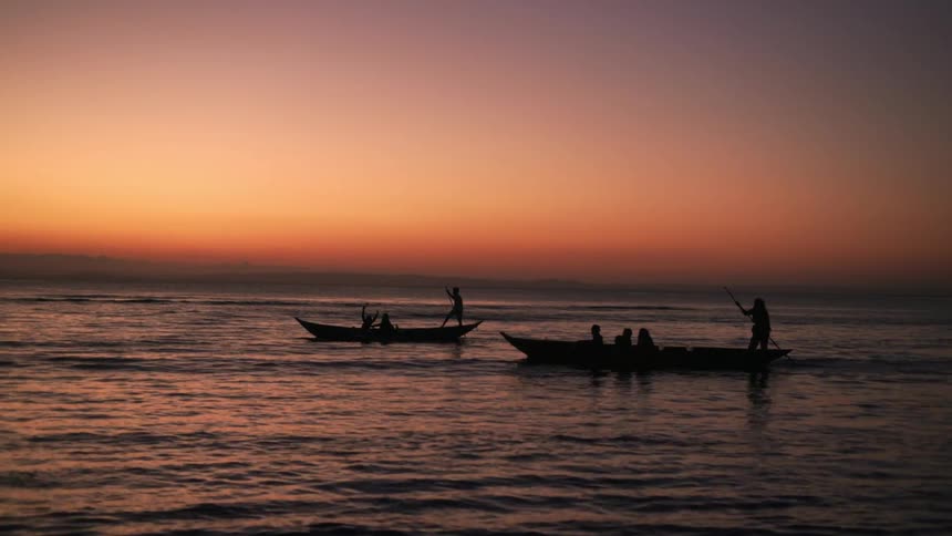 Silhouette of people inside two canoes on calm sea with a reddish dawn sky