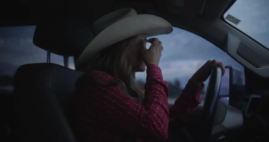A blonde woman in a cowboy hat is driving a car