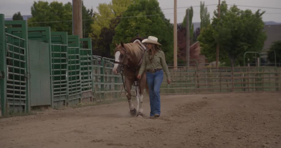 A western style dressed woman in a cowboy hat walks with a horse in an outdoor rodeo arena next to the start gates
