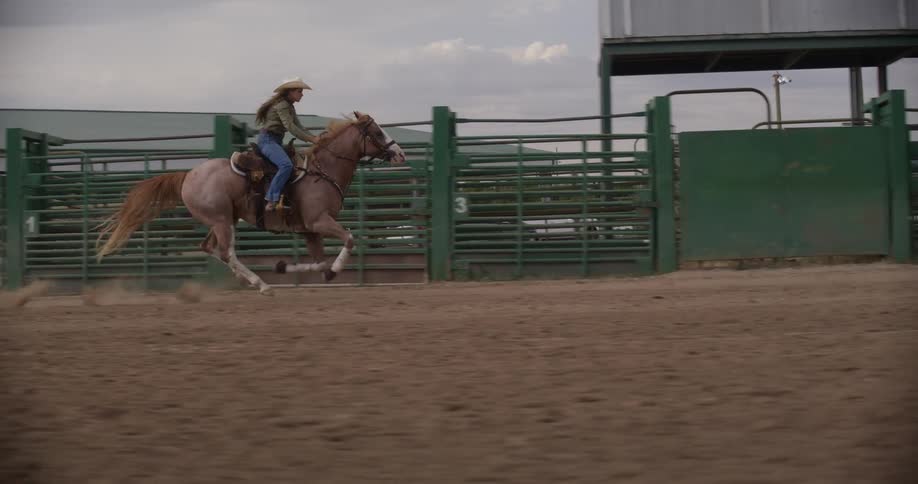 A woman on a horse wearing a cowboy hat riding very fast in a ranch
