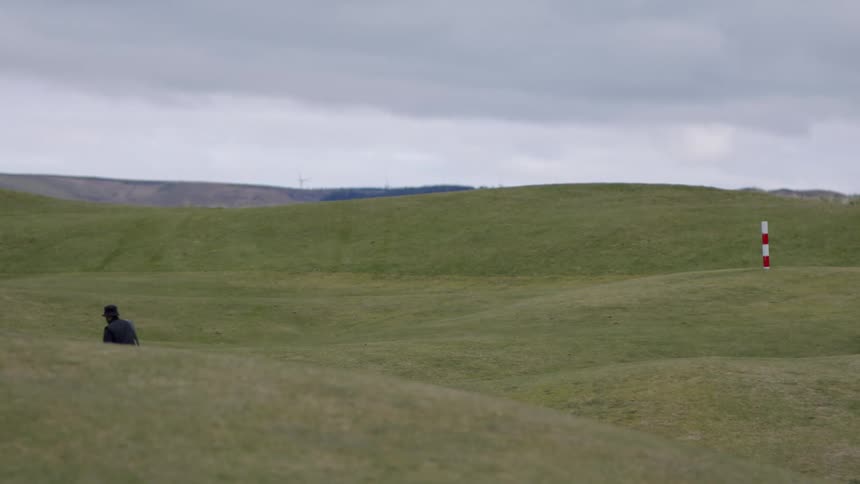 A man walking on a golf course on a cloudy day raises their right arm