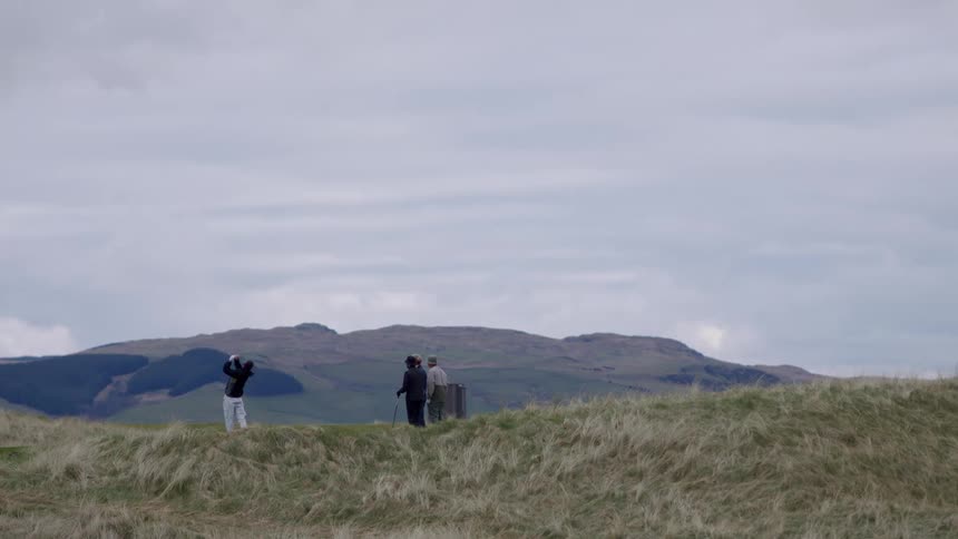 Three golfers stand still while a fourth golfer swings a club on a remote golf course