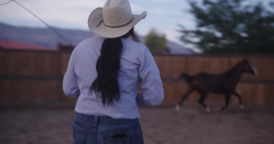 Lady controls the movement of a horse with a whip in a fenced area during the day