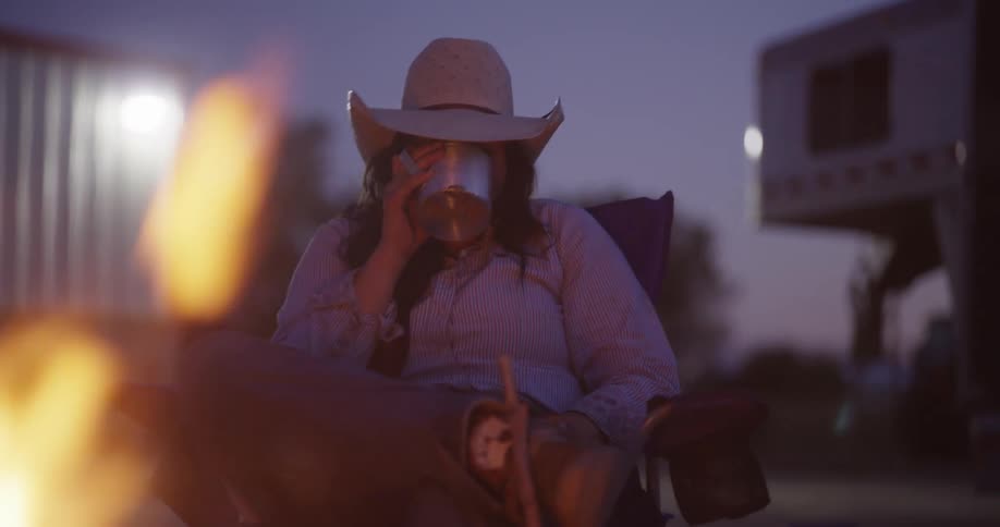 A woman sitting by a camp fire wearing a cowboy hat and drinking coffee from a cup in the evening