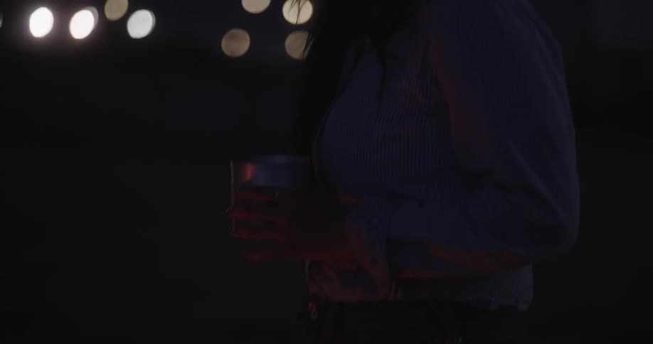 View of a woman with a cowboy hat drinking something from a metal mug at night