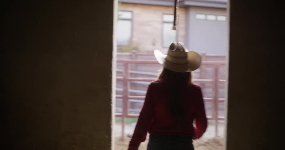 Lady wearing a cowboy hat walks out of a barn during the day