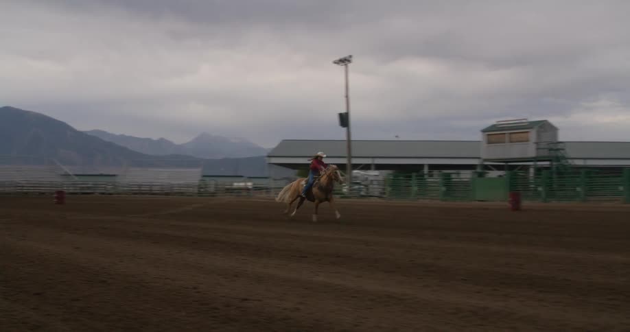A woman races a horse down a dirt track at an empty rodeo stadium under a cloudy sky