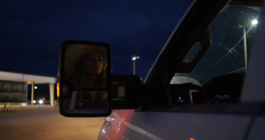 A woman in a cowboy hats reflection shows in the sideview mirror as she looks around while driving into a lit up rodeo parking lot at night