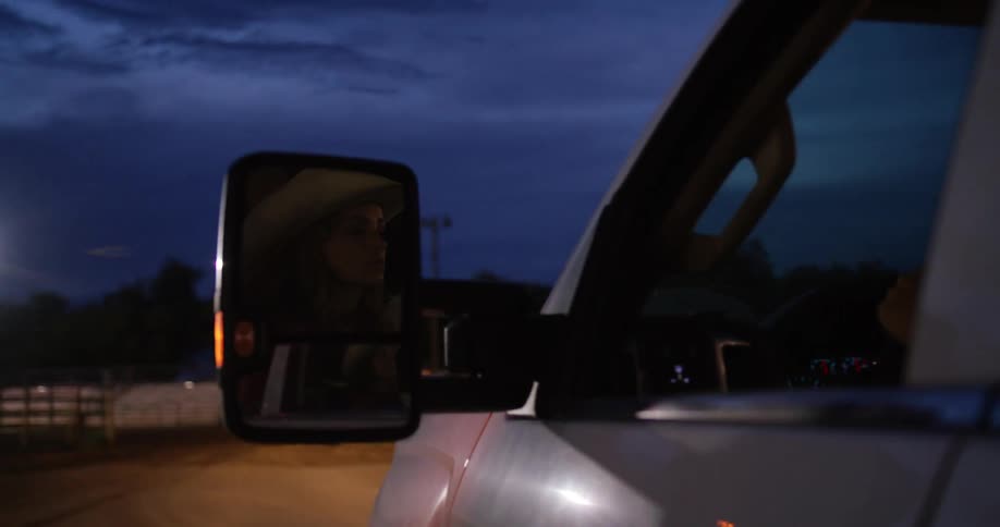 A reflection of a woman with cowboy hat in a cars rearview mirror