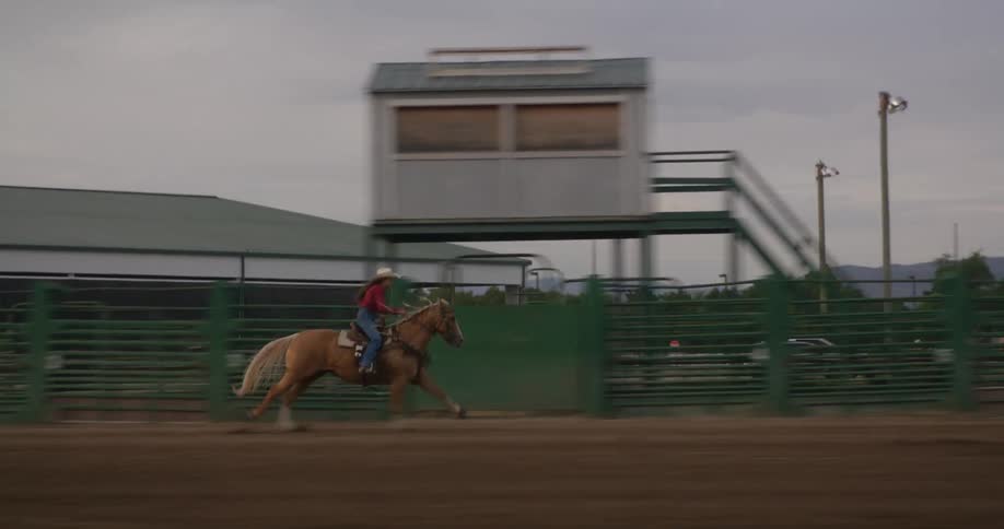 A horse with a female rider runs quickly past an announcer stand kicking up sandy dirt behind it inside the pen at an outdoor rodeo arena at dusk