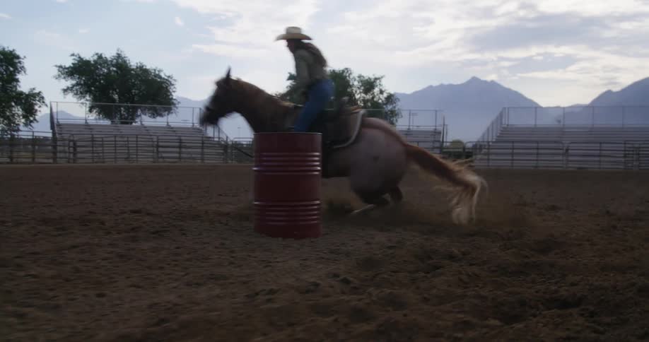 Cowgirl training with a horse and circling a red barrel in the middle of a rodeo arena in the morning