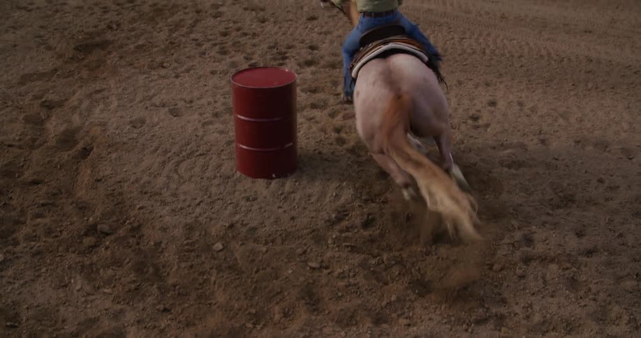 Woman on top of a horse spinning a metal barrel at high speed in a sandy field