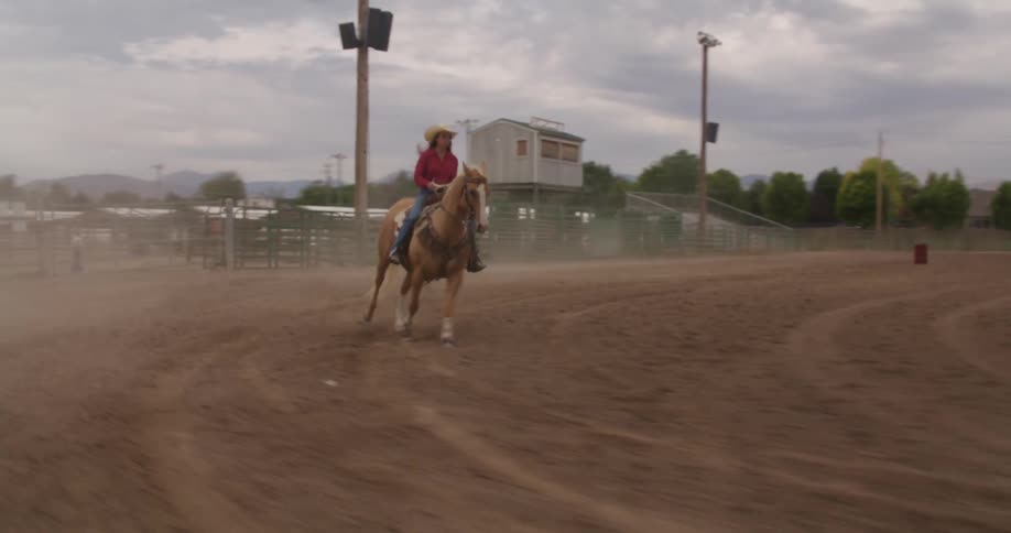 A blonde woman in a hat is riding a horse on a ranch with bleachers on the sides