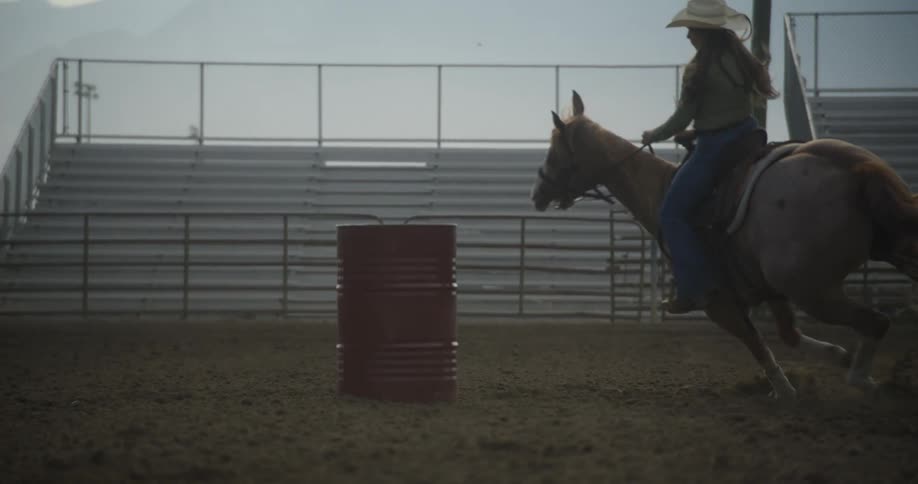 Woman riding a horse around a metal gallon in the middle of a rodeo field