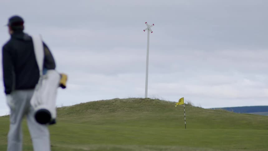 A golfer carrying a white golf bag on his shoulder walks past the hole at the bottom of a hill as the yellow flag blows in the wind under a cloudy sky near a tall red and white x boundary marker