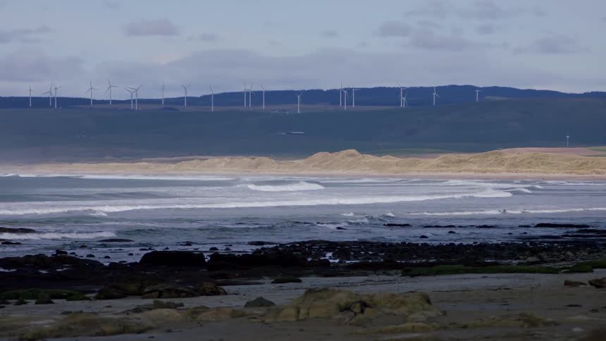 Waves roll into shore as wind turbines spin on a hill in the distance