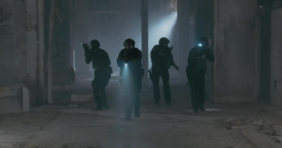 A swat team of four move together through an empty falling down building past rubble moving their guns flashlights around looking as they move forward