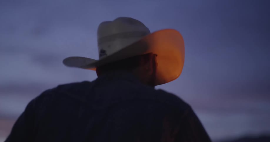 A man wearing a cowboy hat opens a beer bottle and sits by the camp fire at dusk