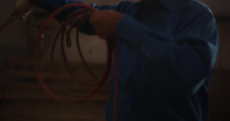 A cowboy standing in a barn throws a rope after swinging it for a while