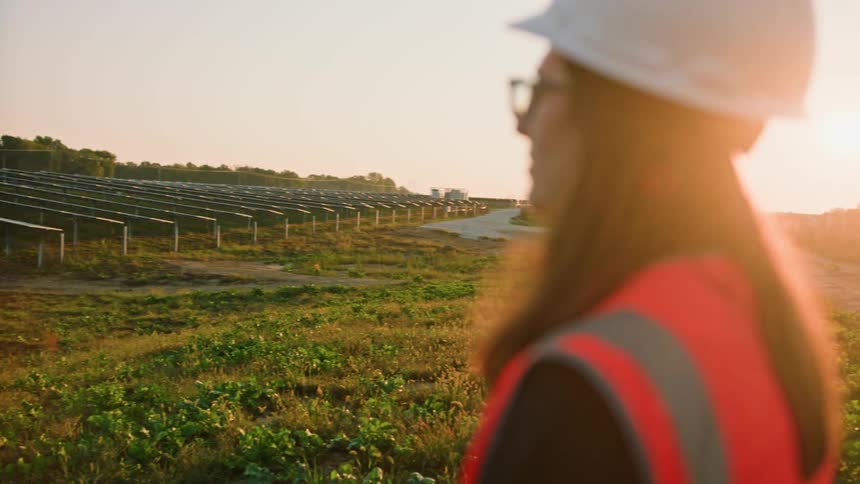 Female worker in safety gear contemplating a solar field at golden hour