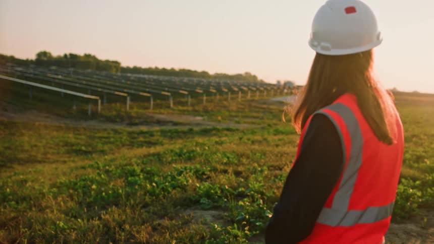 A utility worker walks in a field looking at rows of solar panels on a solar farm at sunset